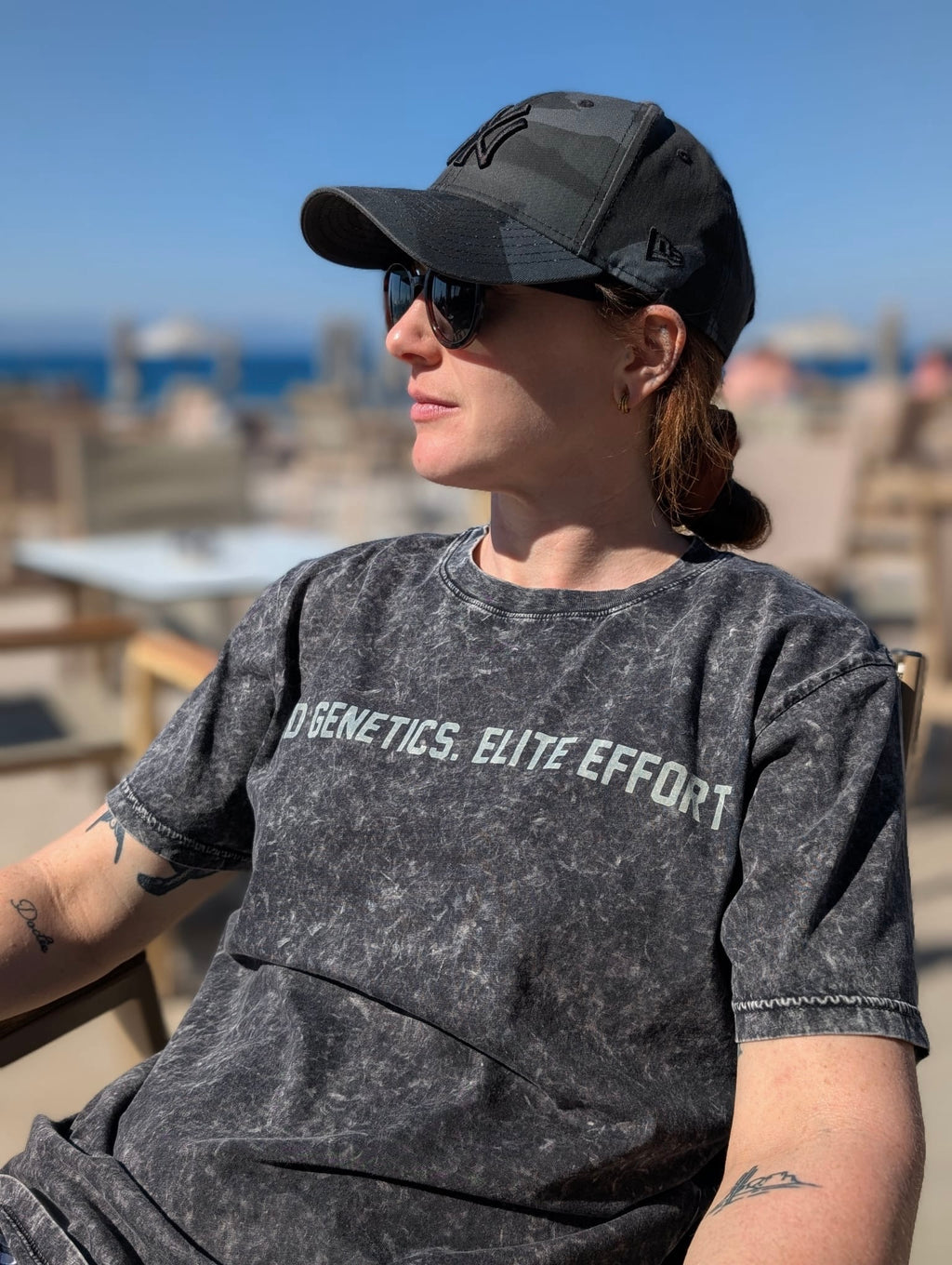 Person wearing a black cap and sunglasses, wearing an acid wash t-shirt sitting outdoors with ocean view