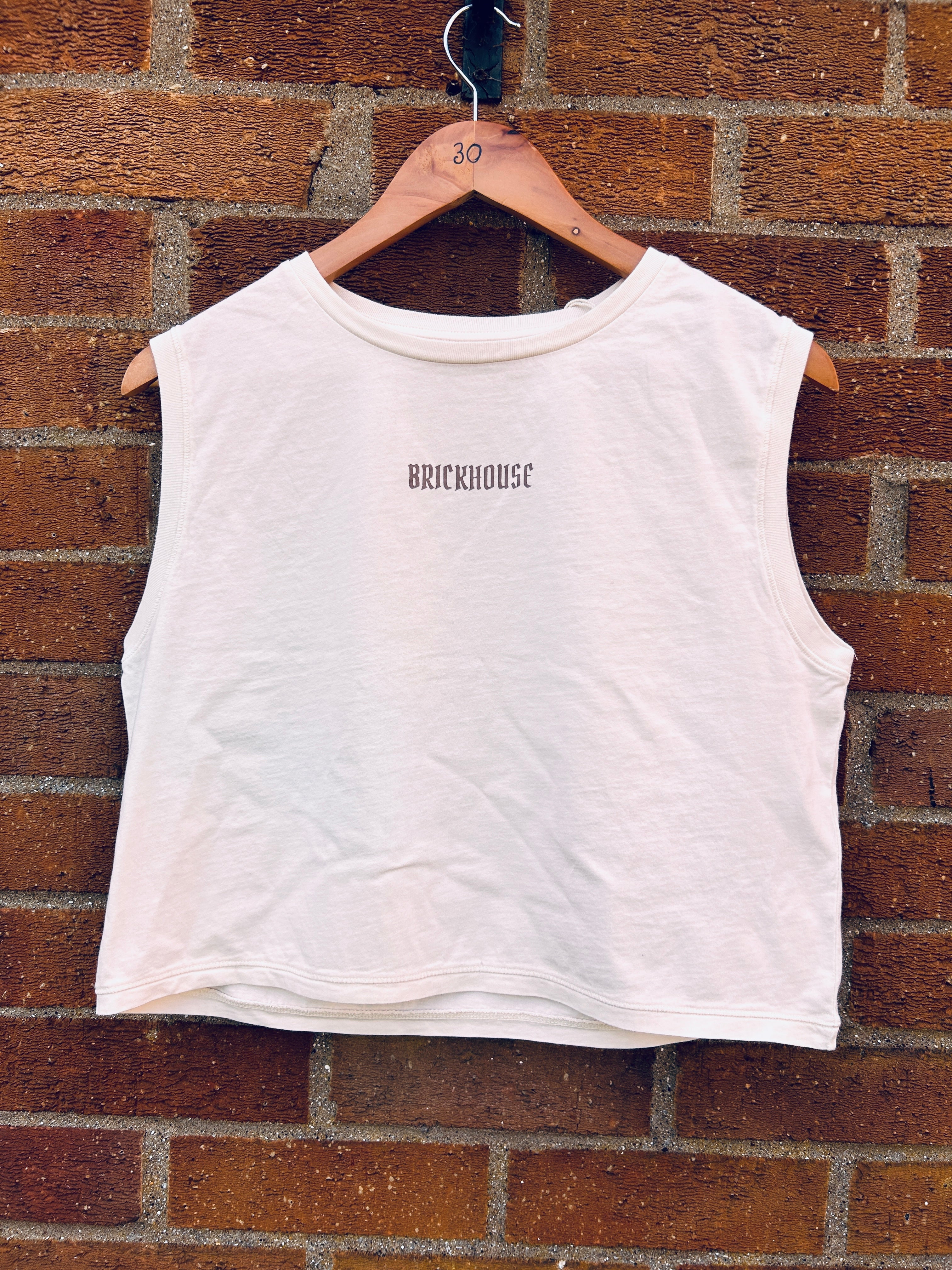 White sleeveless shirt with 'BRICKHOUSE' text on a hanger against a brick wall.