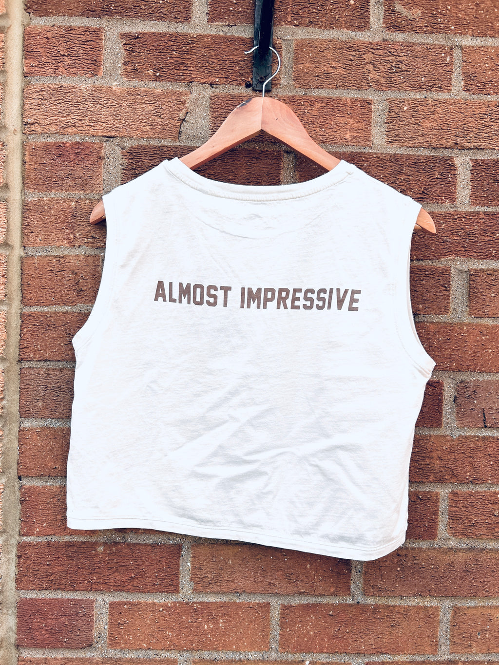 White sleeveless shirt with 'ALMOST IMPRESSIVE' text on a hanger against a brick wall.
