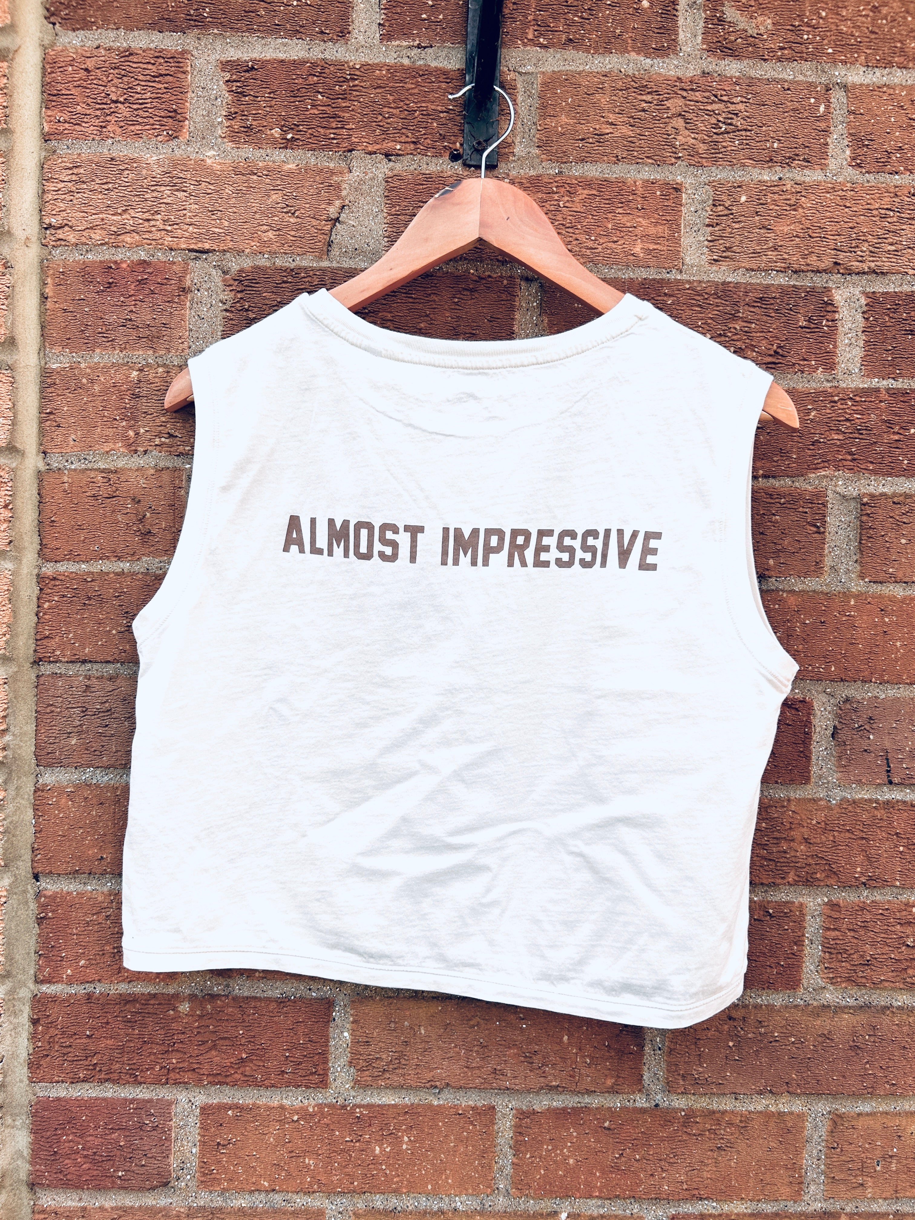 White sleeveless shirt with 'ALMOST IMPRESSIVE' text on a hanger against a brick wall.