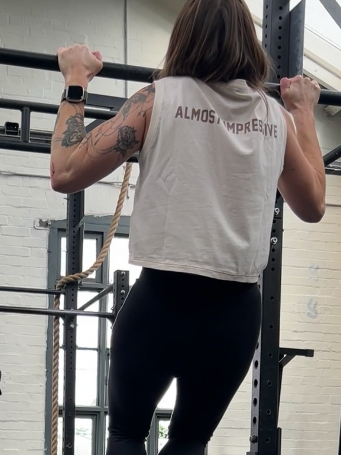 Woman wearing a sleeveless shirt with 'ALMOST IMPRESSIVE' text, performing pull-ups.