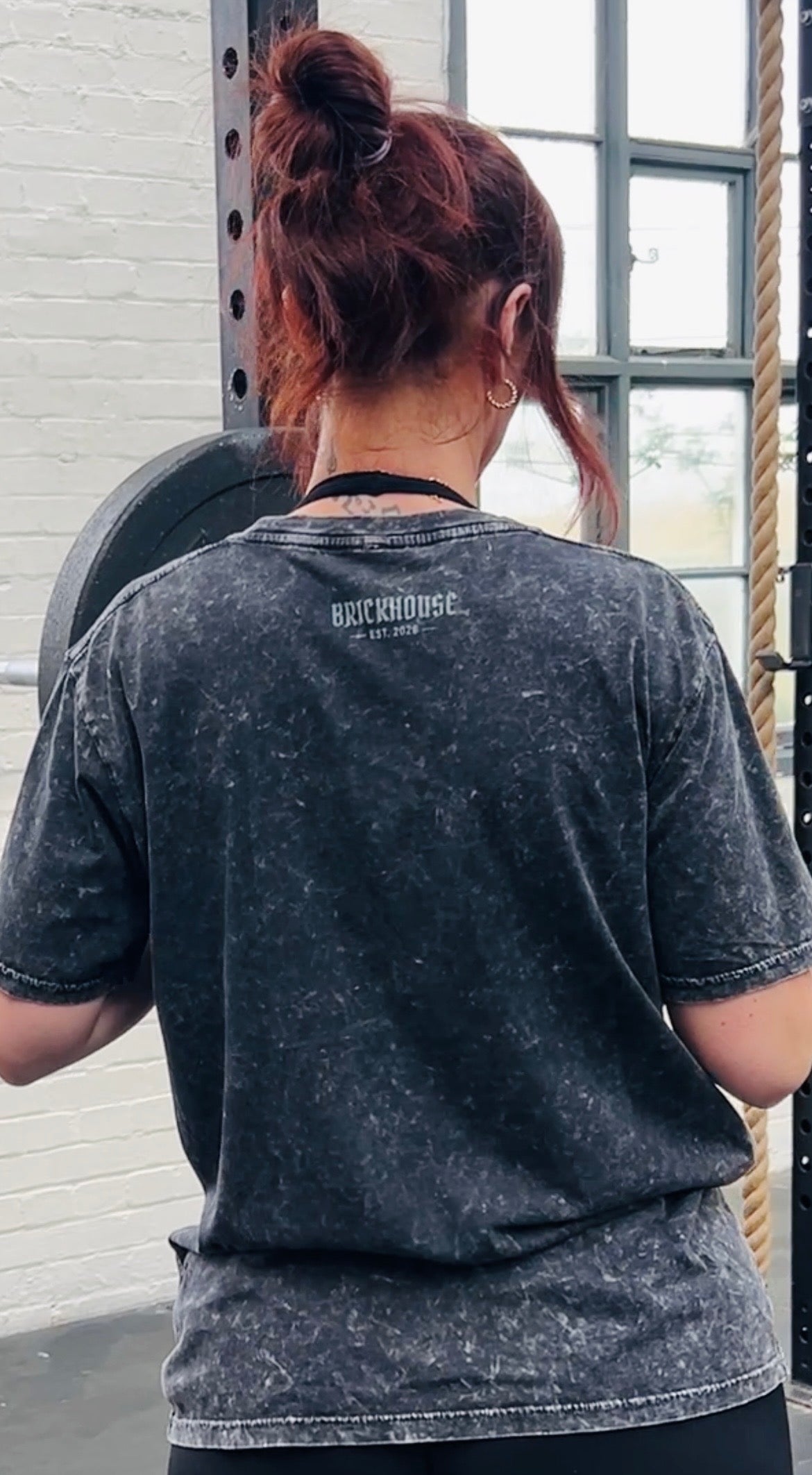 Woman wearing an acid washed black t-shirt with 'BRICKHOUSE' logo text at a gym.