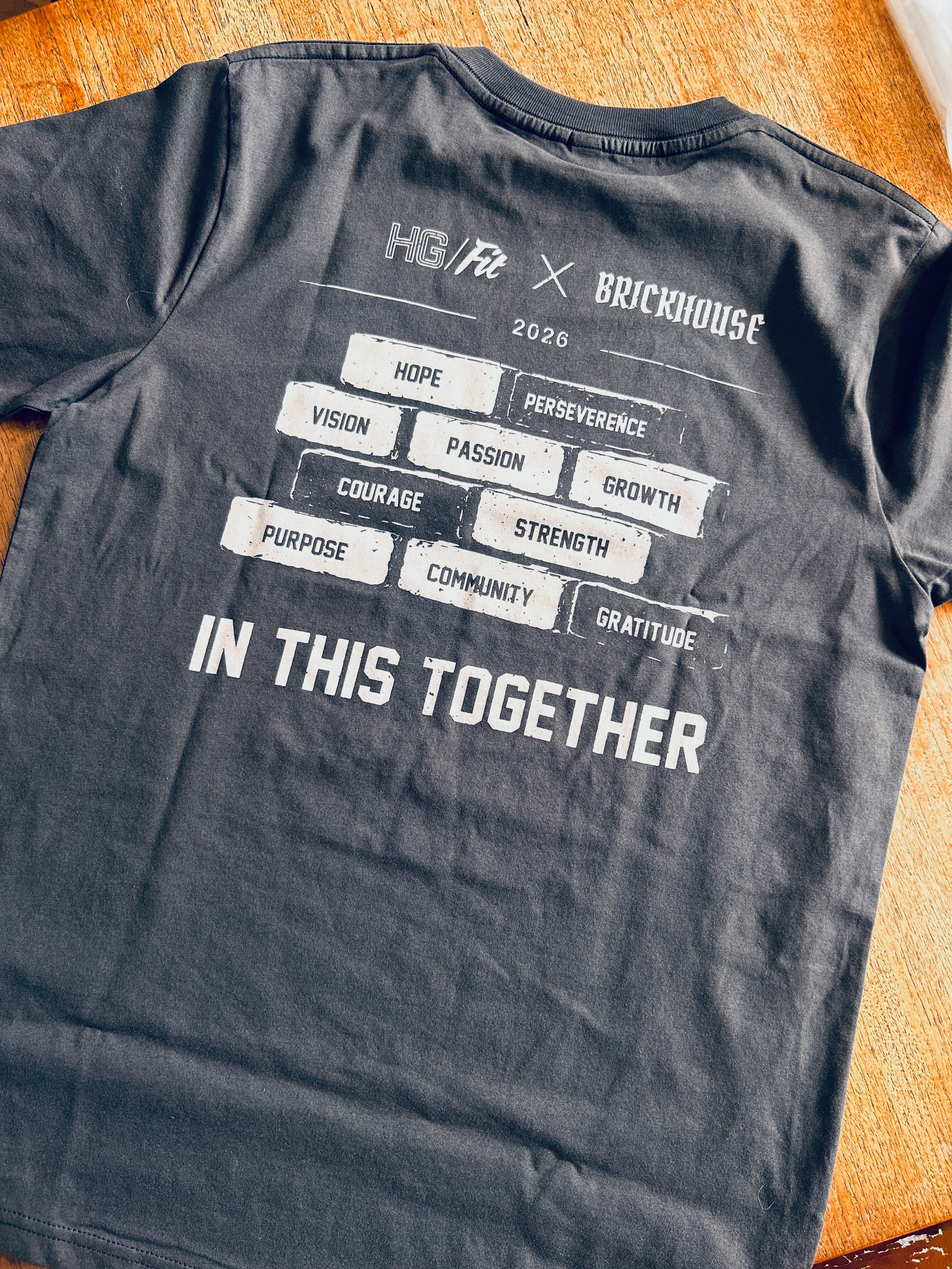 Gray t-shirt with motivational text and HG Fit and Brickhouse logos on a wooden surface