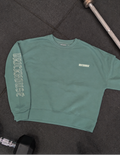 A sage green crewneck sweatshirt laid flat on a dark gym floor, with 'BRICKHOUSE' printed vertically on one sleeve and a small matching logo embroidered on the left chest. A barbell and dumbbells are partially visible around it.