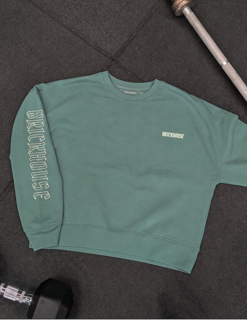 A sage green crewneck sweatshirt laid flat on a dark gym floor, with 'BRICKHOUSE' printed vertically on one sleeve and a small matching logo embroidered on the left chest. A barbell and dumbbells are partially visible around it.