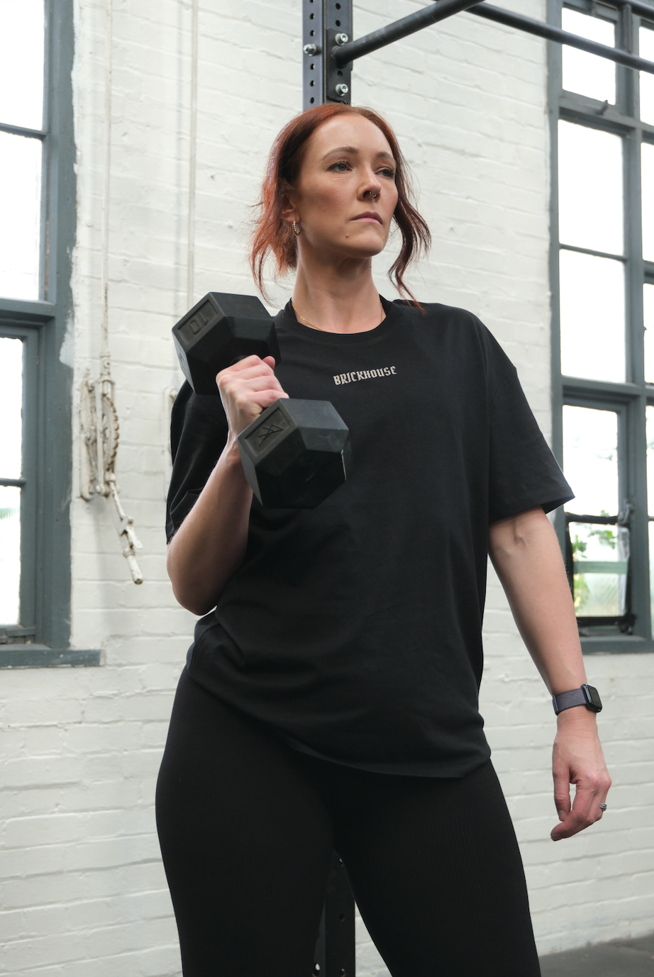 Woman lifting a dumbbell in a gym setting wearing a black t-shirt with BRICKHOUSE text logo.